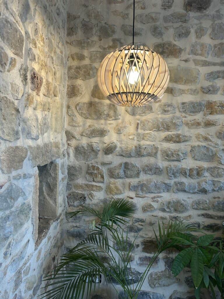 Une lampe en bois suspendue devant un mur de pierre et au dessus d'une plante