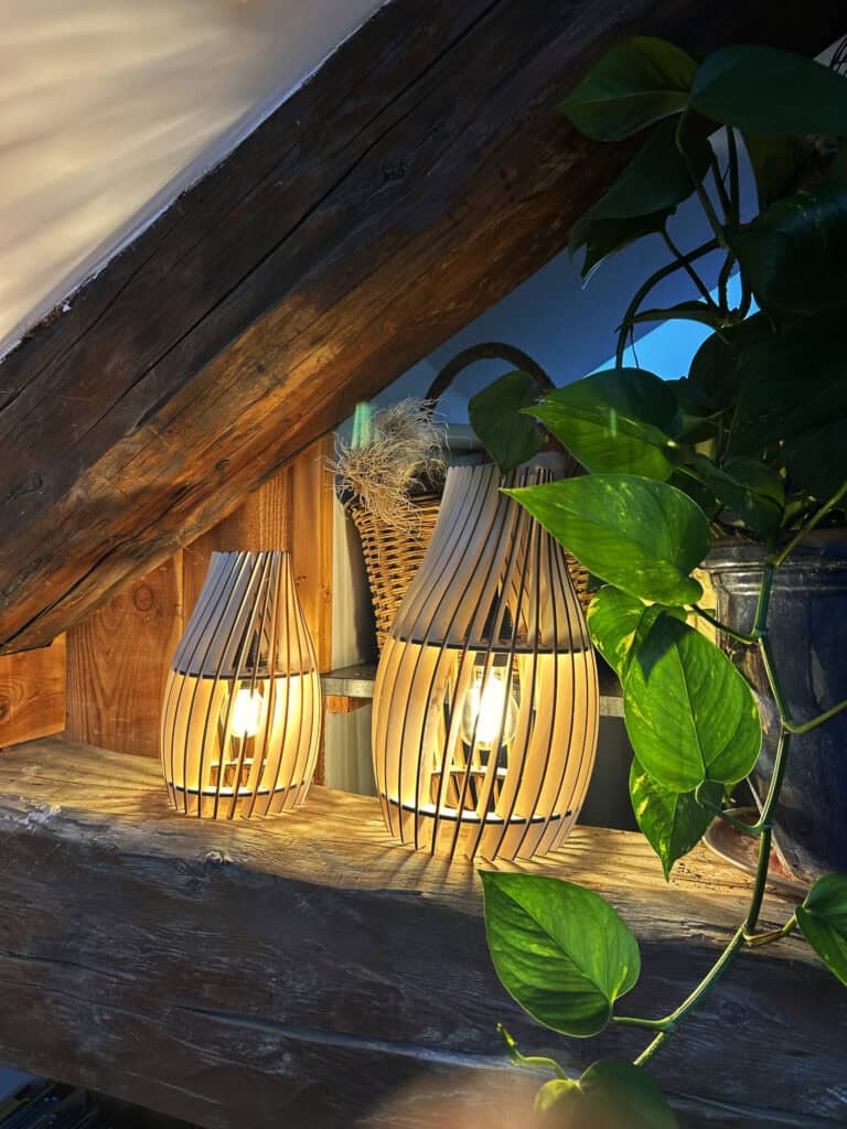 Deux lampes de table en forme de goutte en bois découpé Atelier Sapi, allumées sur une étagère en bois brut sous des poutres apparentes, avec plantes vertes