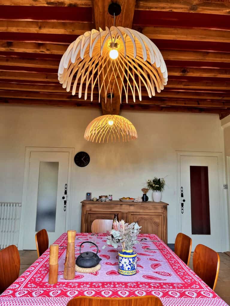 Deux suspensions en bois découpé modèle Dôme Atelier Sapi suspendues au-dessus d'une grande table à manger avec nappe provençale rouge, sous des poutres en bois peintes en rouge