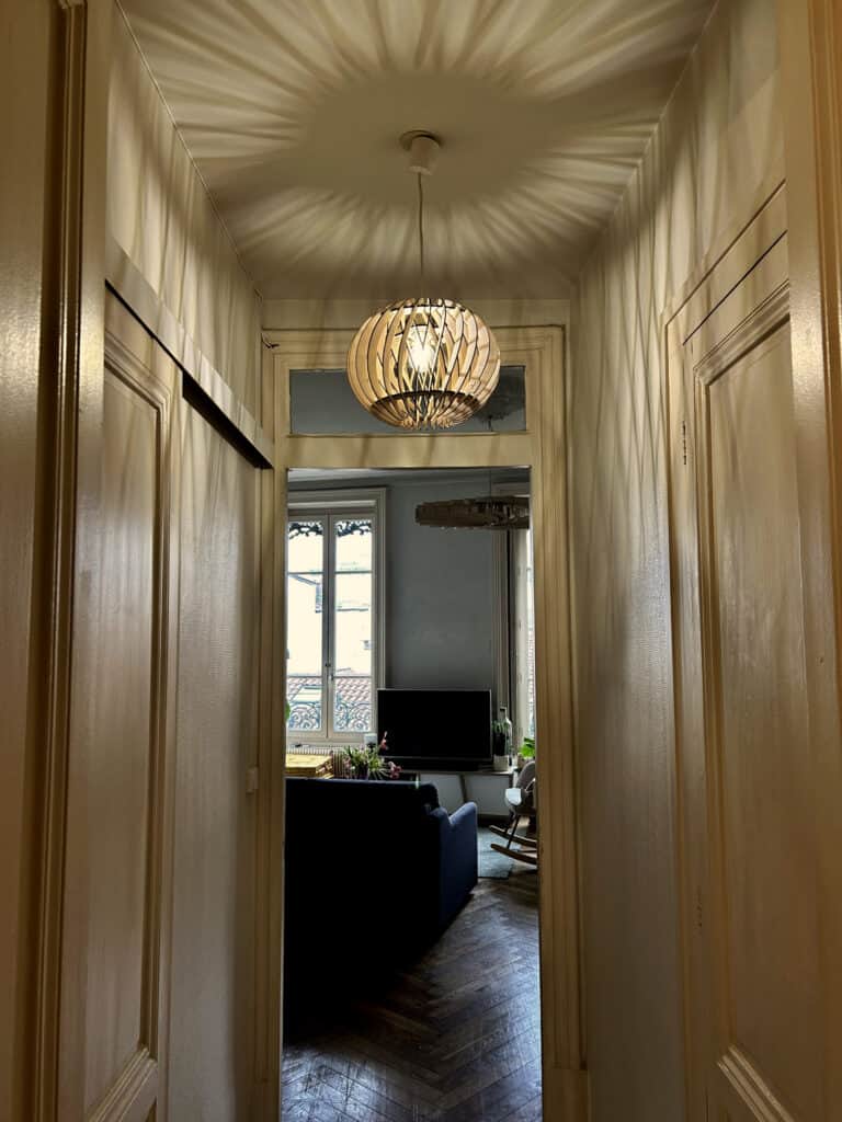 Suspension sphérique en bois découpé Atelier Sapi dans un couloir haussmannien avec portes en bois, projetant des ombres rayonnantes sur le plafond