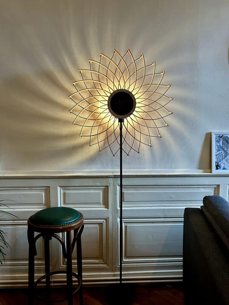 Lampadaire sur pied modèle Soleil en bois découpé Atelier Sapi allumé devant un mur blanc avec lambris et moulures, tabouret bistrot vert et canapé gris