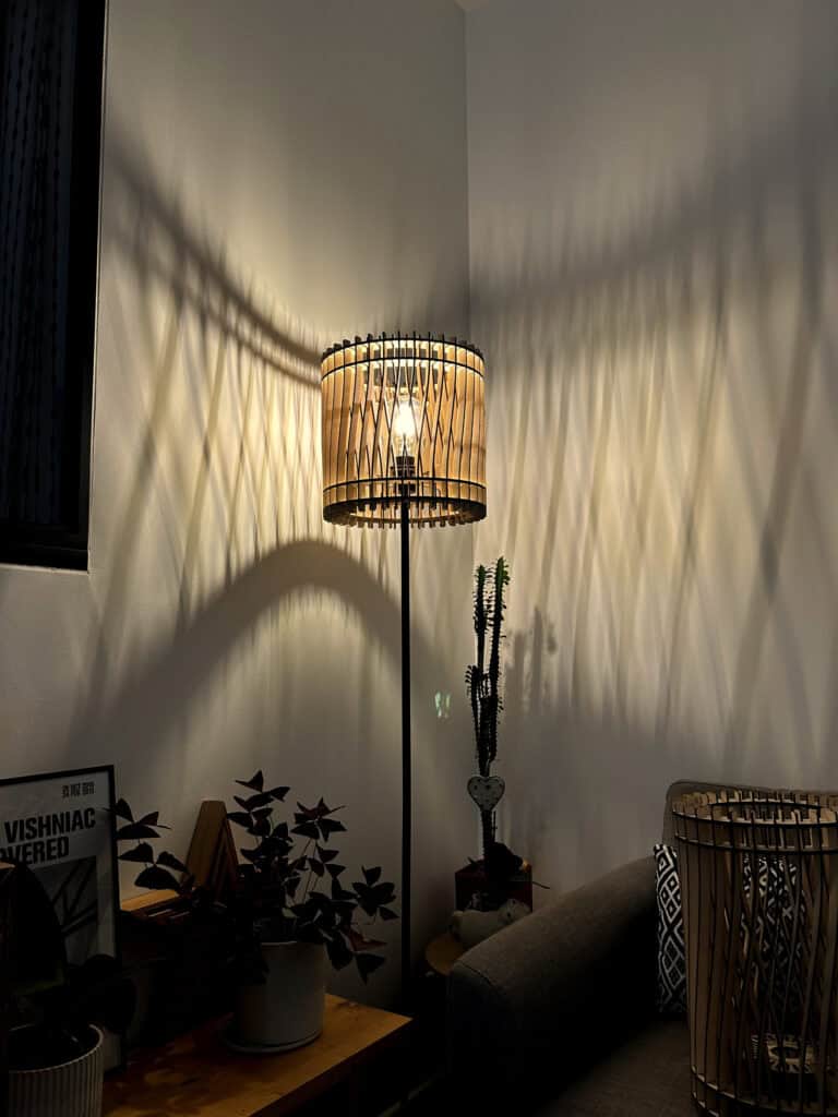 Gros plan sur la suspension en bois découpé modèle Chapeau Chinois Atelier Sapi allumée le soir, ombres rayonnantes sur mur blanc avec moulures, canapé sombre en bas de cadre