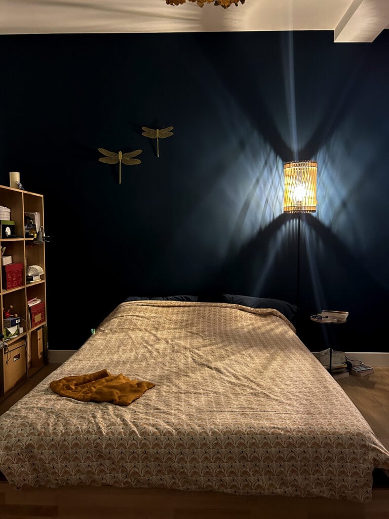Lampe de table cylindrique en bois Atelier Sapi posée sur une table de chevet, projetant des ombres en éventail sur un mur bleu nuit avec décorations libellules dorées, lit double au premier plan