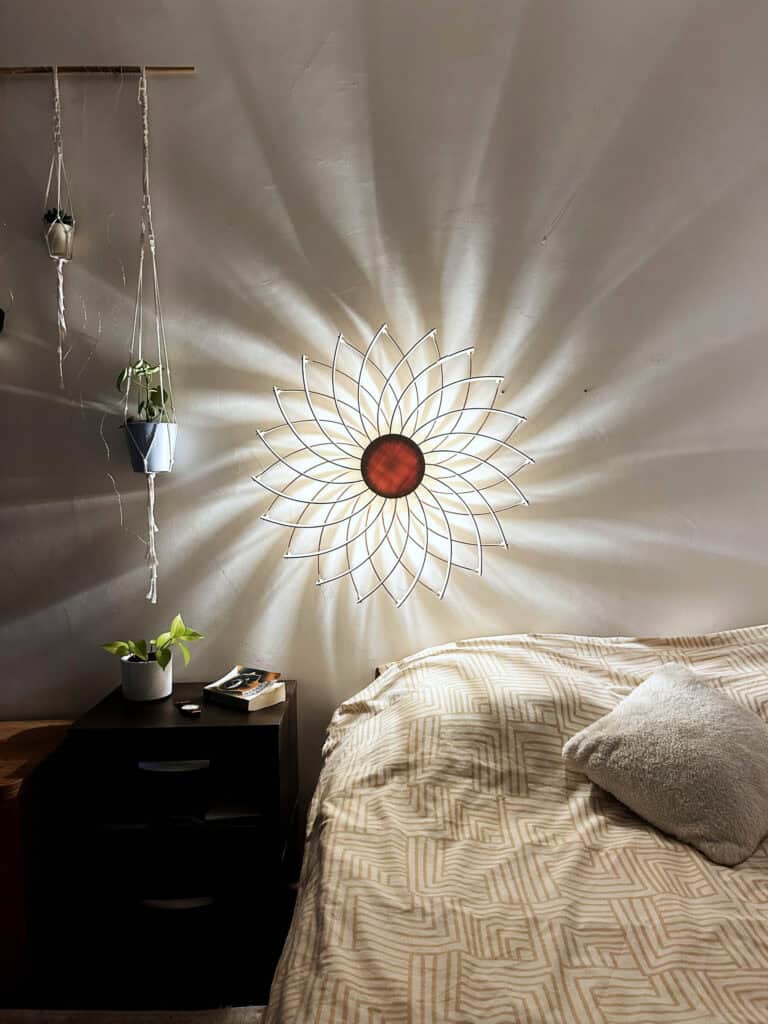Applique murale modèle Soleil en bois découpé Atelier Sapi fixée au mur au-dessus d'un lit, projetant des ombres rayonnantes sur mur blanc, plantes suspendues en macramé et table de nuit noire