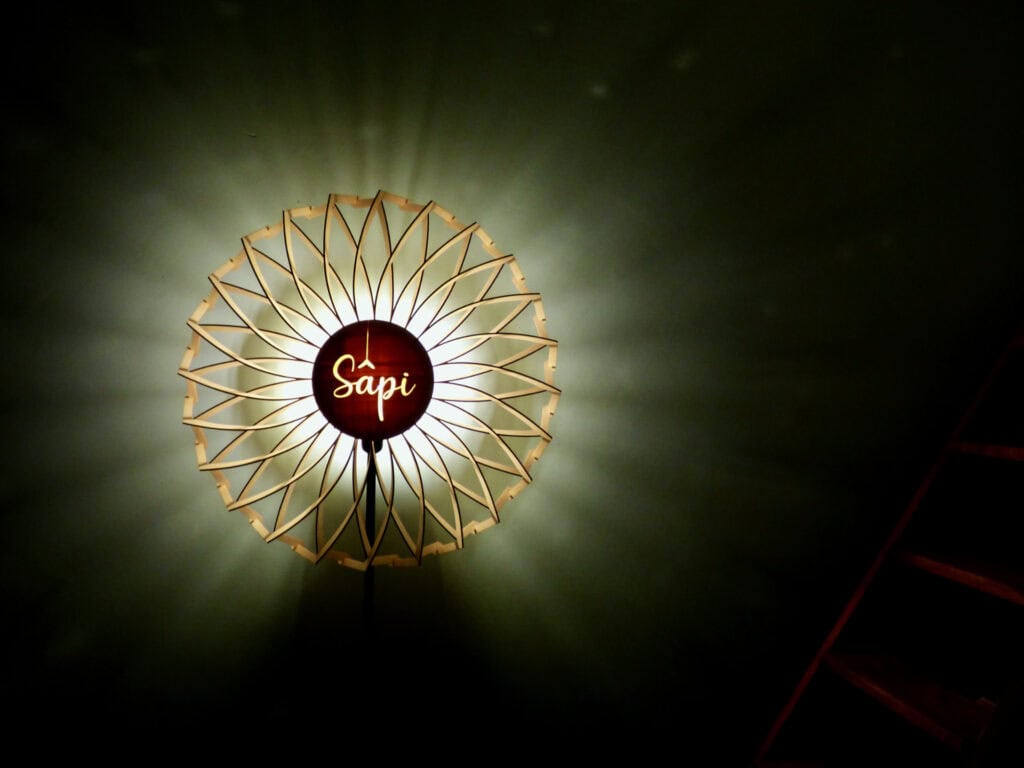 Lampadaire Signature modèle Marguerite Atelier Sapi photographié sur fond noir, abat-jour mandala en bois découpé à double rangée de pétales droits avec médaillon central gravé Sâpi, halo lumineux doré