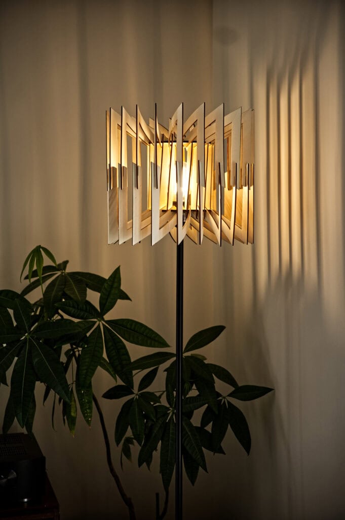Le lampadaire géométrique Léon allumé contrastant avec les grandes feuilles d'une plante verte d'intérieur.