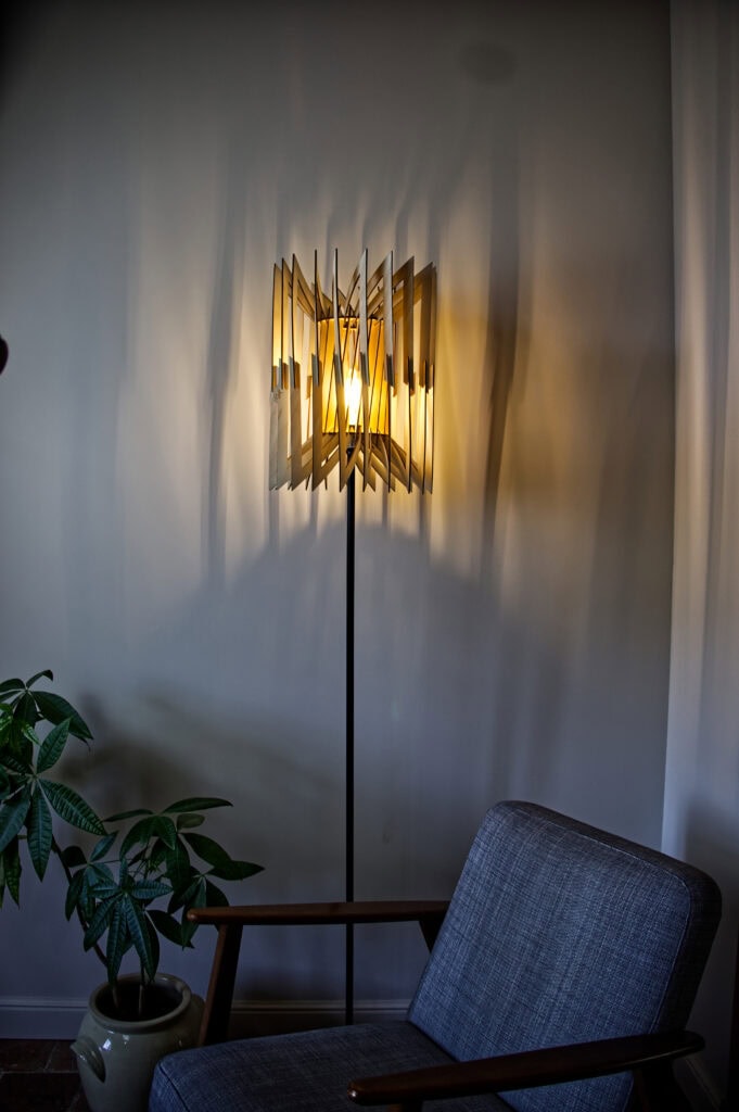 Lampadaire Léon allumé à côté d'un fauteuil gris moderne, projetant des ombres graphiques sur le mur.